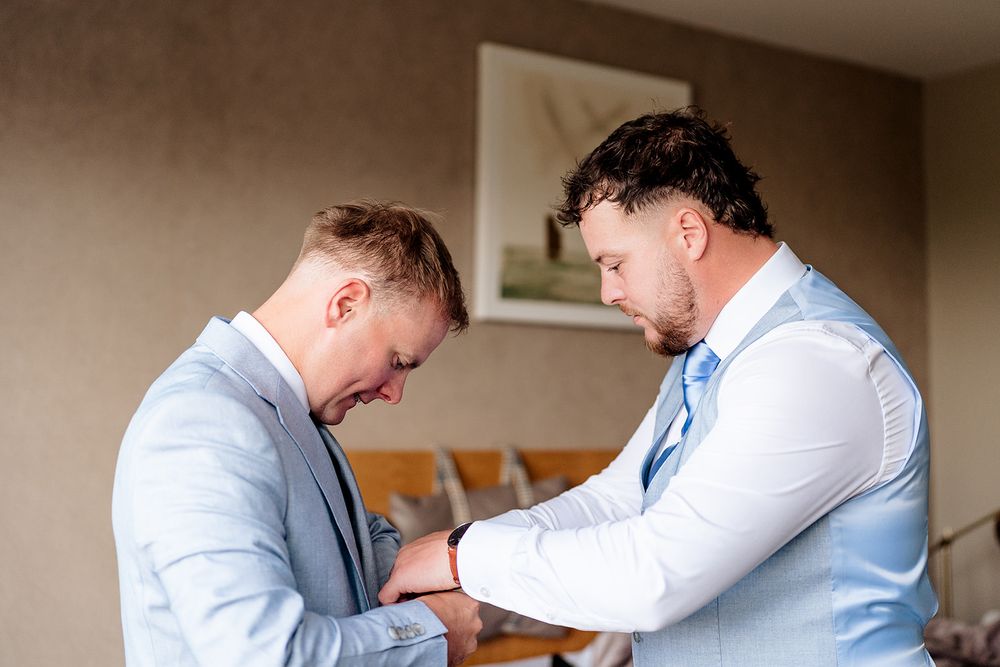 Two men in light blue dress shirts help each other with formal attire preparation.