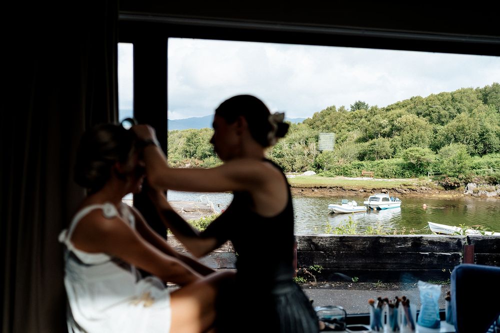 Two people in dark attire prepare for a wedding at a scenic waterfront venue with a boat in the background.