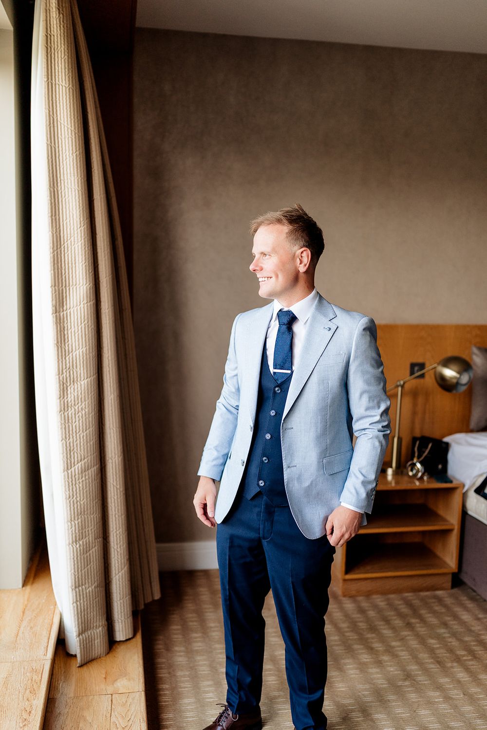 A person in a light gray blazer and navy suit stands near a curtained window in a hotel room.