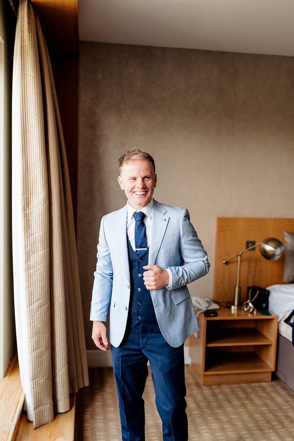 A person wearing a light blue blazer and navy blue tie stands in a hotel room with curtains and wooden furniture.