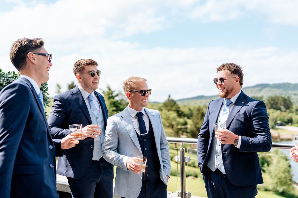 A group of well-dressed gentlemen in suits and sunglasses share drinks and conversation on an outdoor terrace.