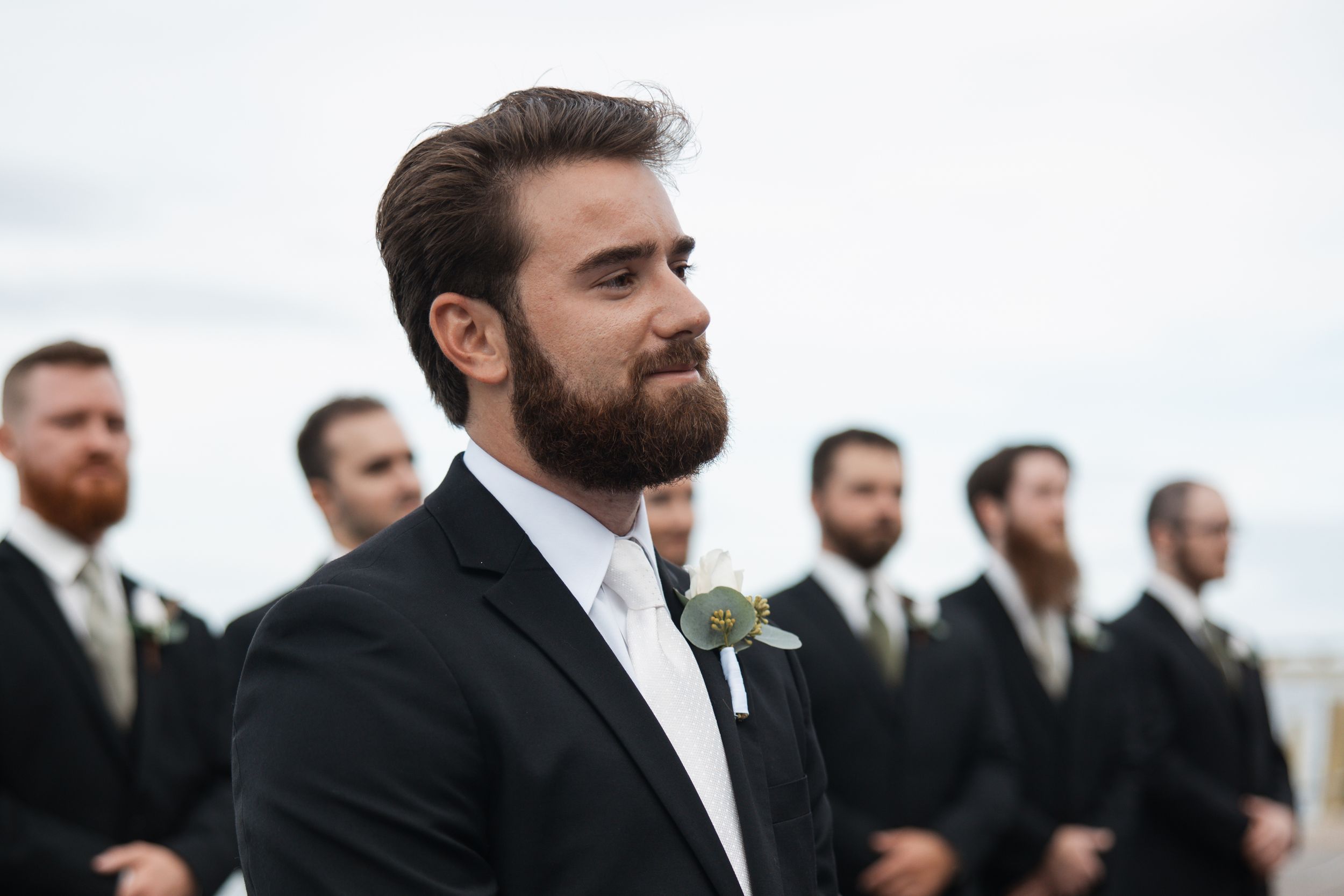 Groomsmen in black suits stand together during an outdoor wedding ceremony with overcast skies.