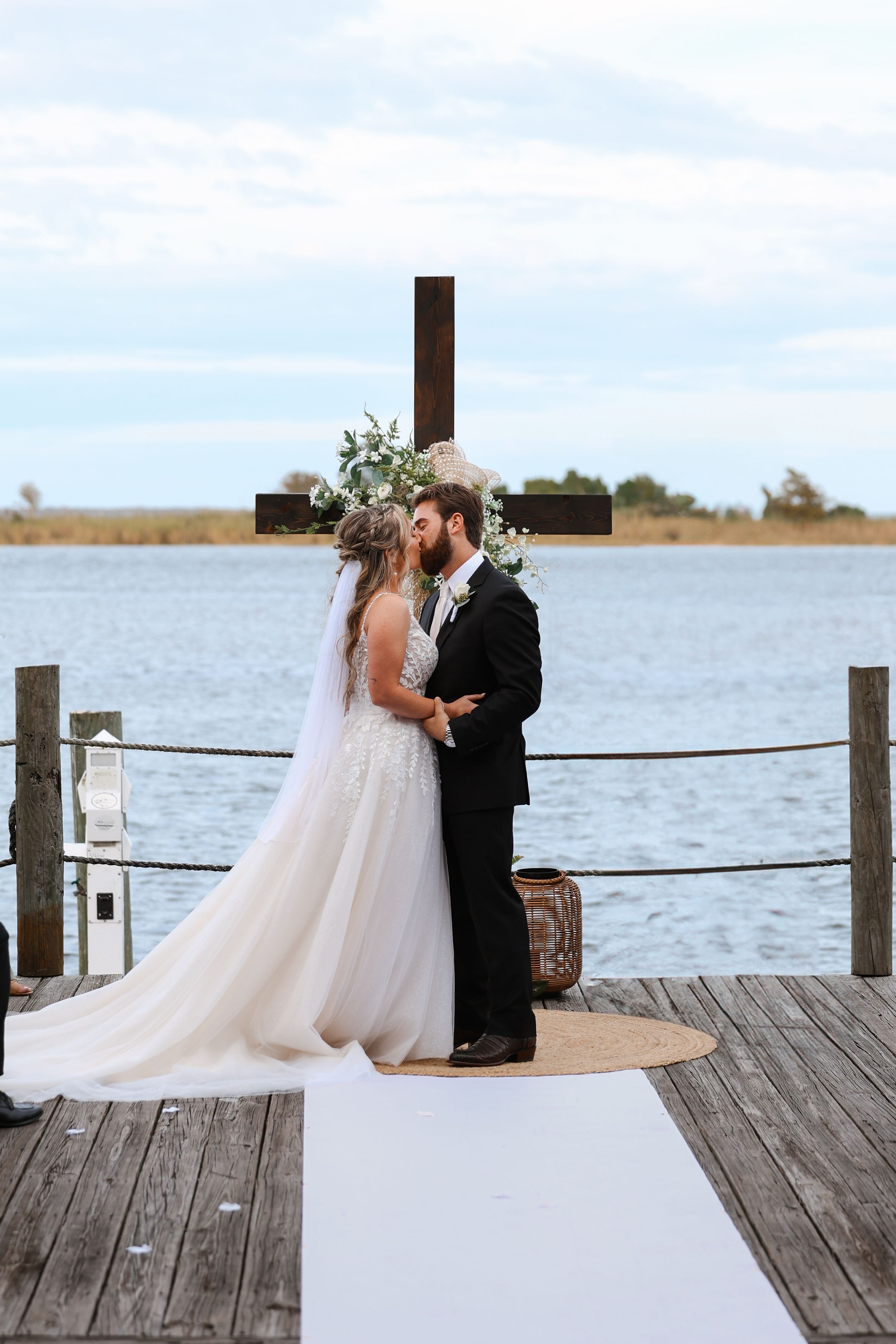 A wedding ceremony takes place on a waterfront dock with a cross in the background on a cloudy day.