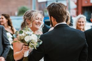 A joyful wedding moment captured outdoors with white floral bouquet against a brick building backdrop.