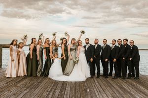 Wedding party poses together for group photos on a wooden pier at sunset with cloudy sky backdrop.