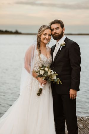 A bride and groom pose together on a dock at sunset, with calm waters in the background.
