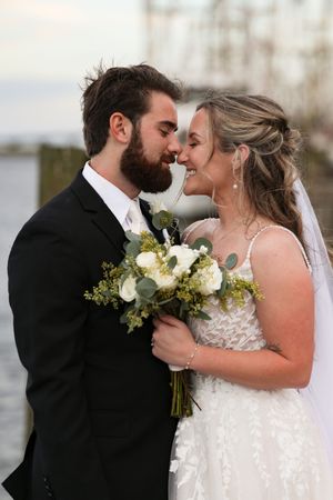 A romantic wedding couple shares an intimate moment while holding a white and green bouquet by the water.