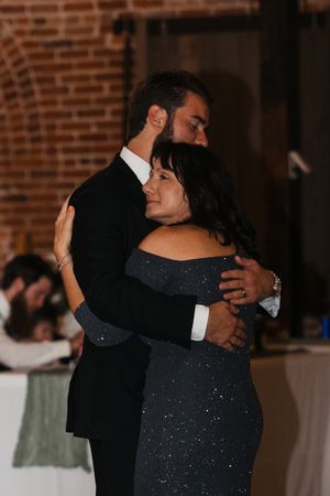 A couple dances together at a wedding reception in a rustic brick-walled venue as they embrace in formal attire.