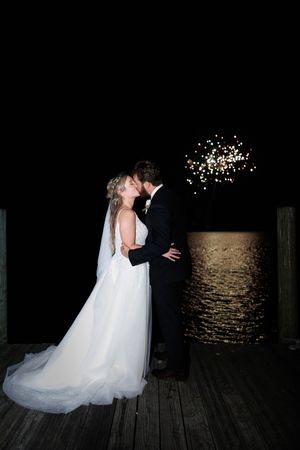 A couple shares romantic moments on a dark wooden dock illuminated by twinkling lights reflecting on calm waters at night.