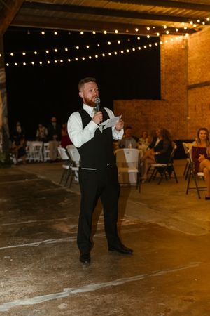 A wedding reception speaker delivers remarks in a rustic venue with string lights overhead.