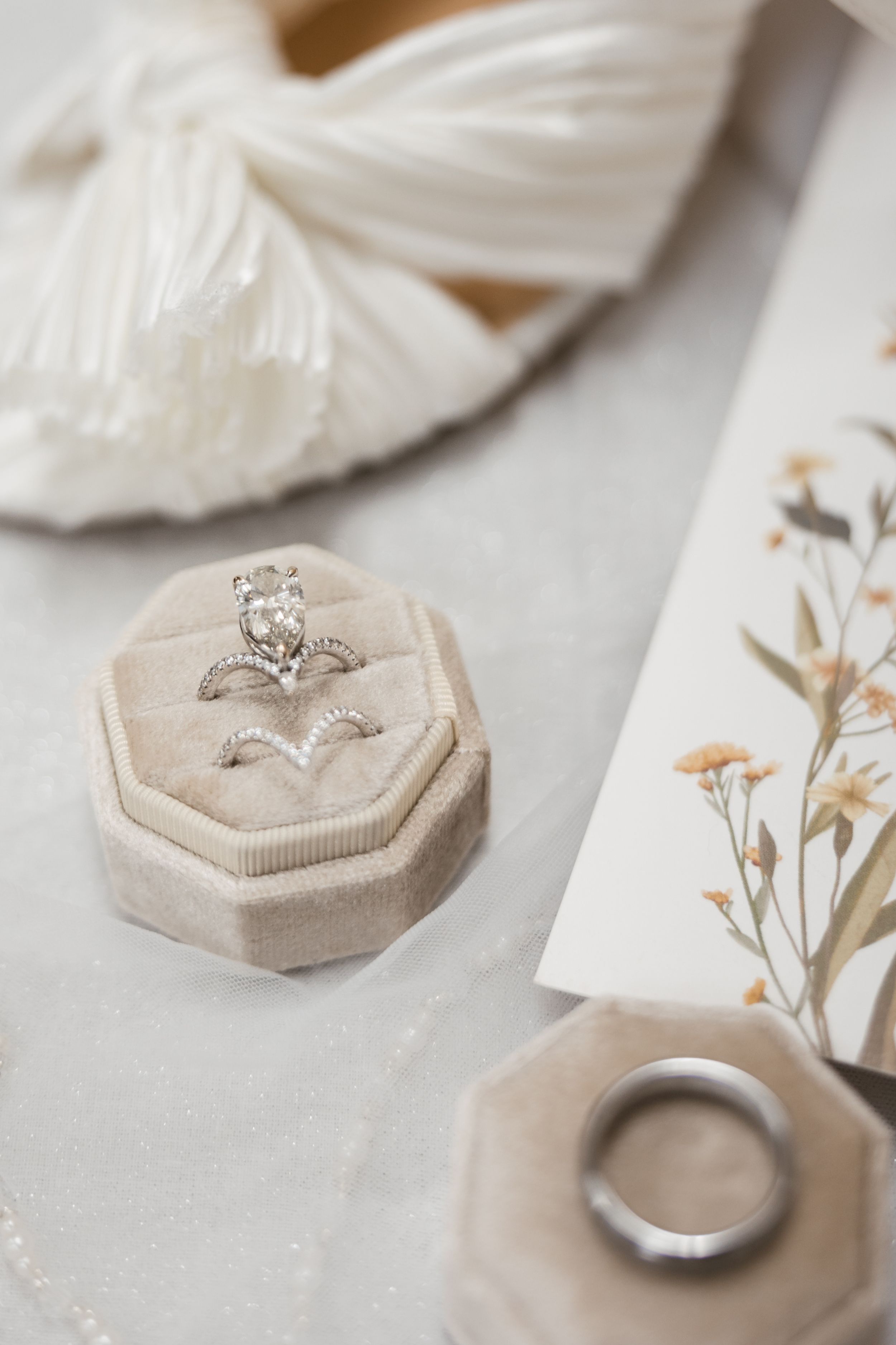 Diamond engagement ring rests in beige velvet octagonal ring box with wedding band and dried flowers nearby.