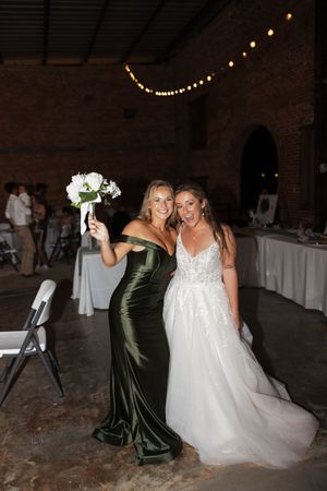 Two women pose together at a wedding reception, one in emerald green velvet and one in a white beaded gown.