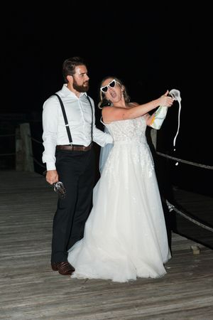 A bride in a white gown and sunglasses poses with her partner in suspenders on a dark wooden deck at night.