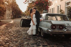 A romantic wedding couple stands beside a classic vintage car on a cobblestone street during autumn.