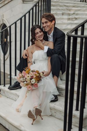 A romantic wedding couple shares candid moments on marble steps with elegant black railings as the bride holds a bouquet.