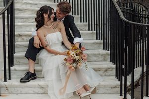 A couple in wedding attire share an intimate moment while sitting on white marble steps with a peach floral bouquet.