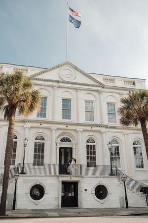 White colonial-style building with arched windows, palm trees, and American flags along the Charleston waterfront.