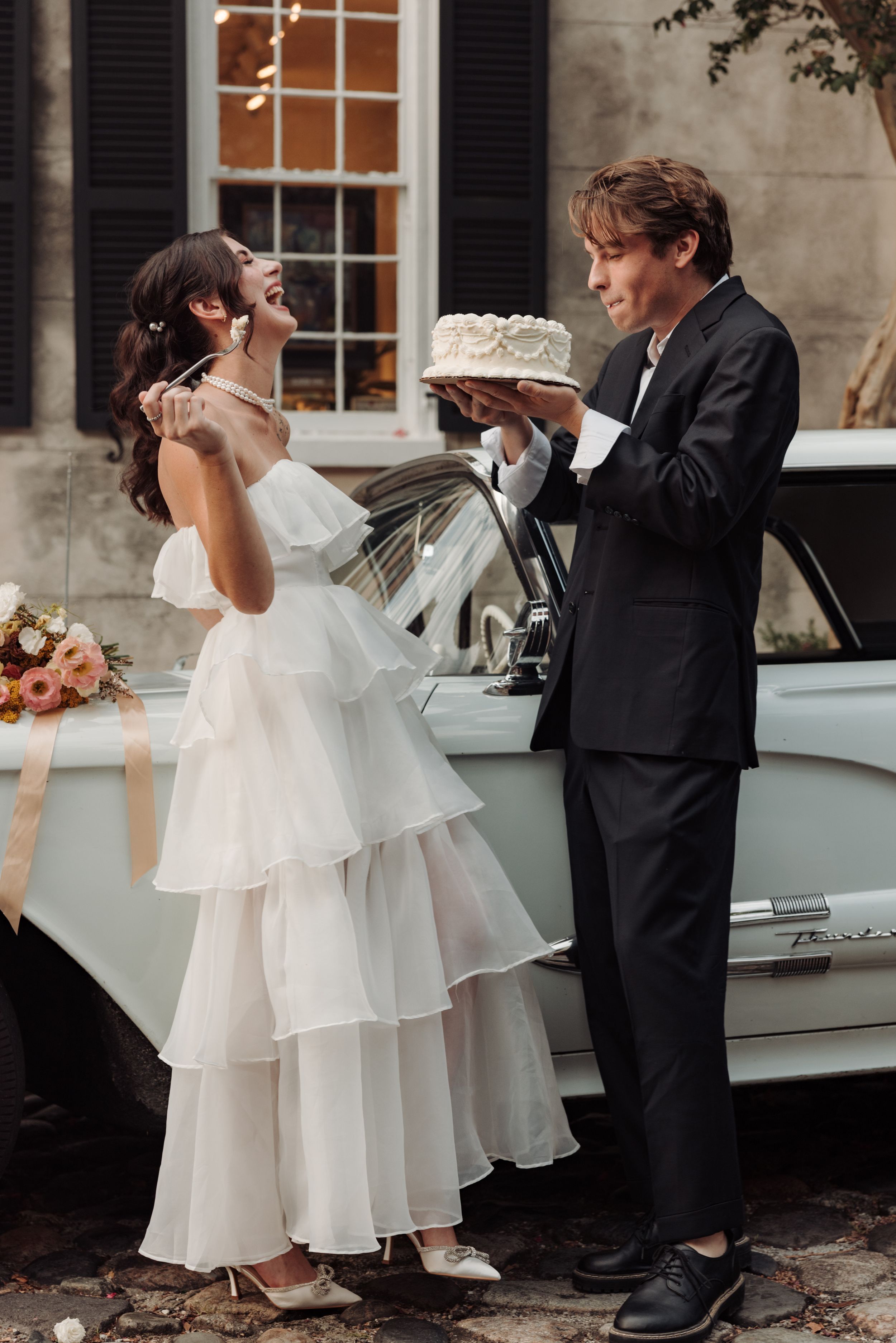 A bride in a white layered dress and groom in black suit dancing playfully by a vintage car during their wedding.