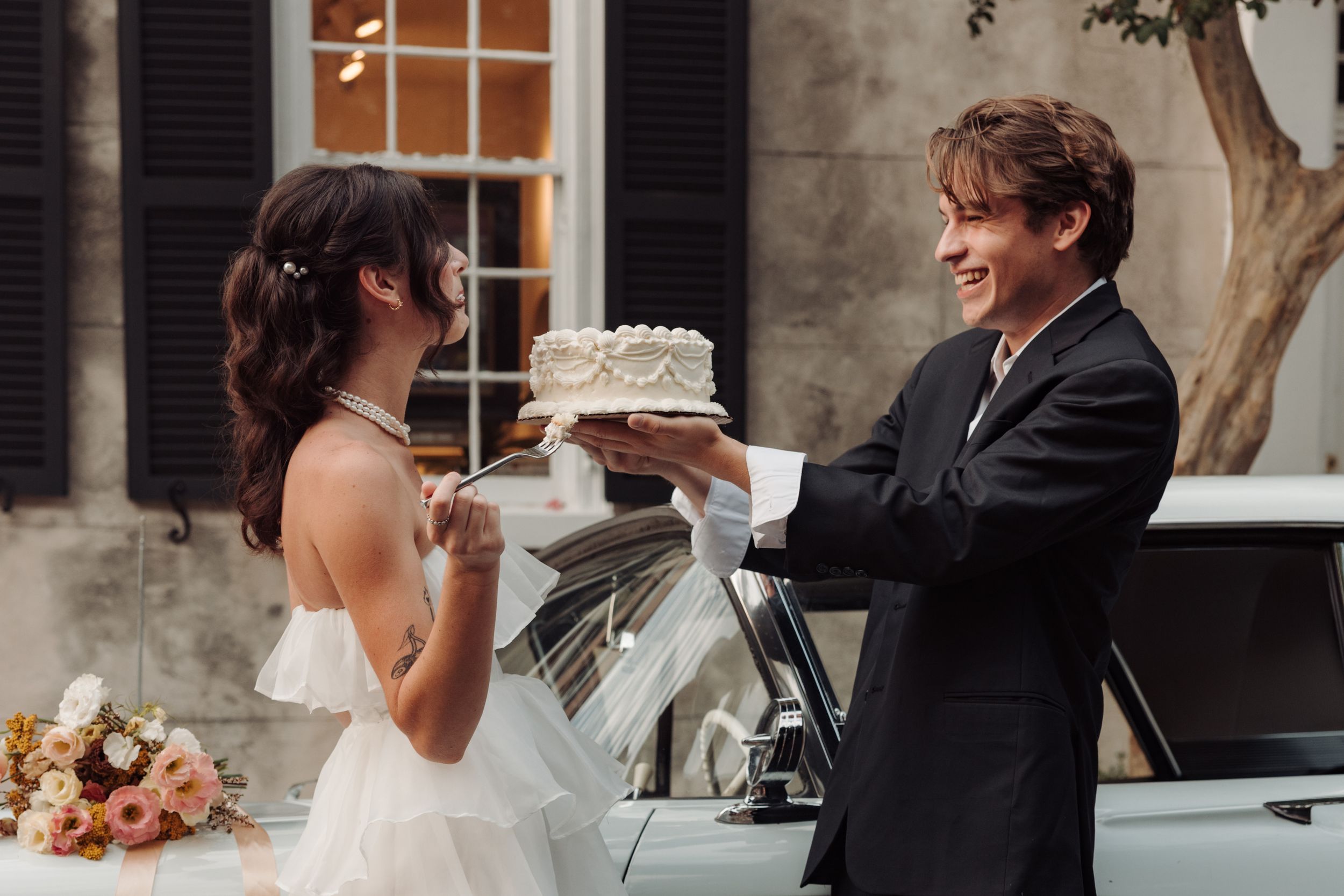 A bride and groom share a playful moment while holding their white wedding cake outdoors near a vintage car.
