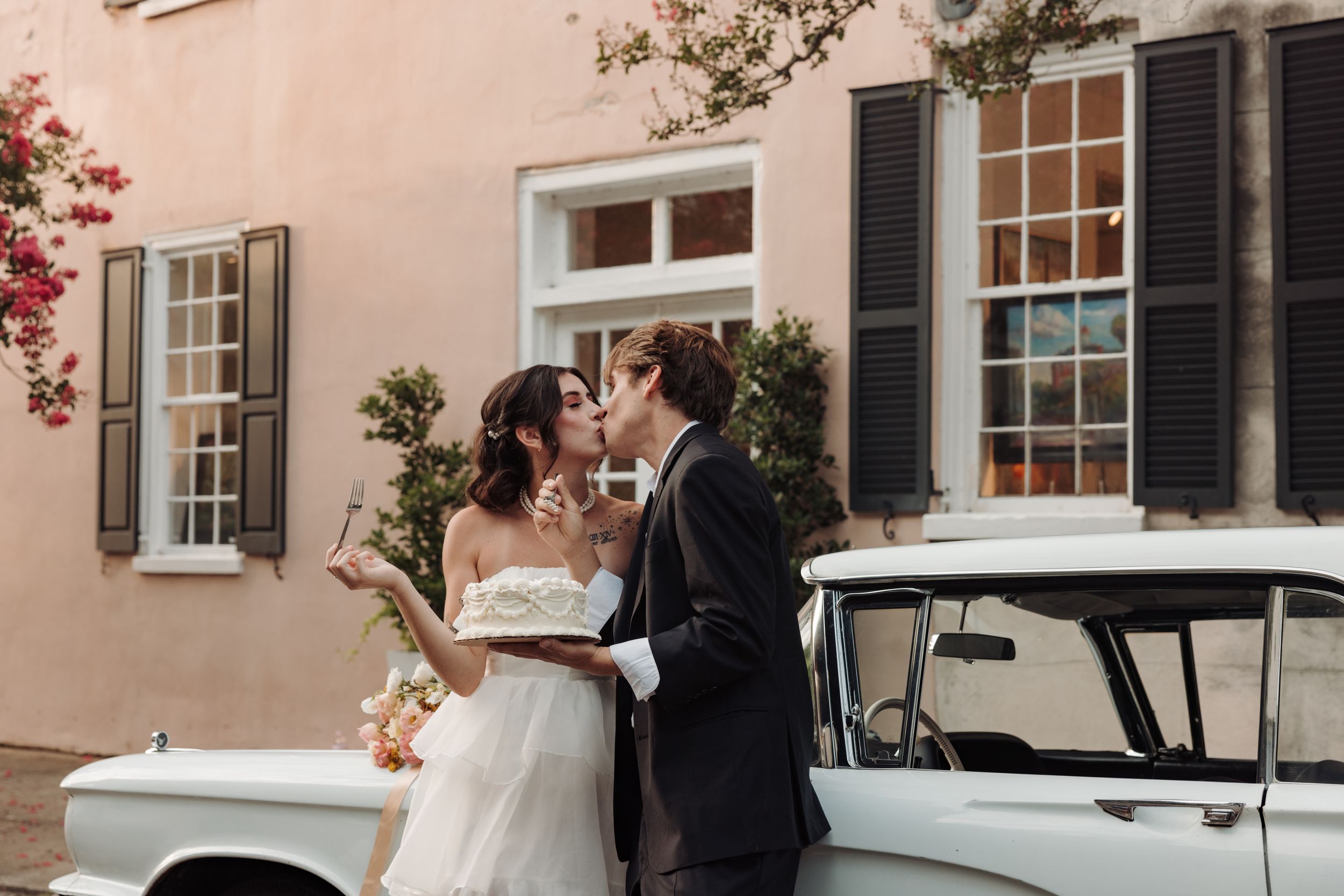 A couple in wedding attire embraces in front of a pink stucco building with a vintage white car parked nearby.