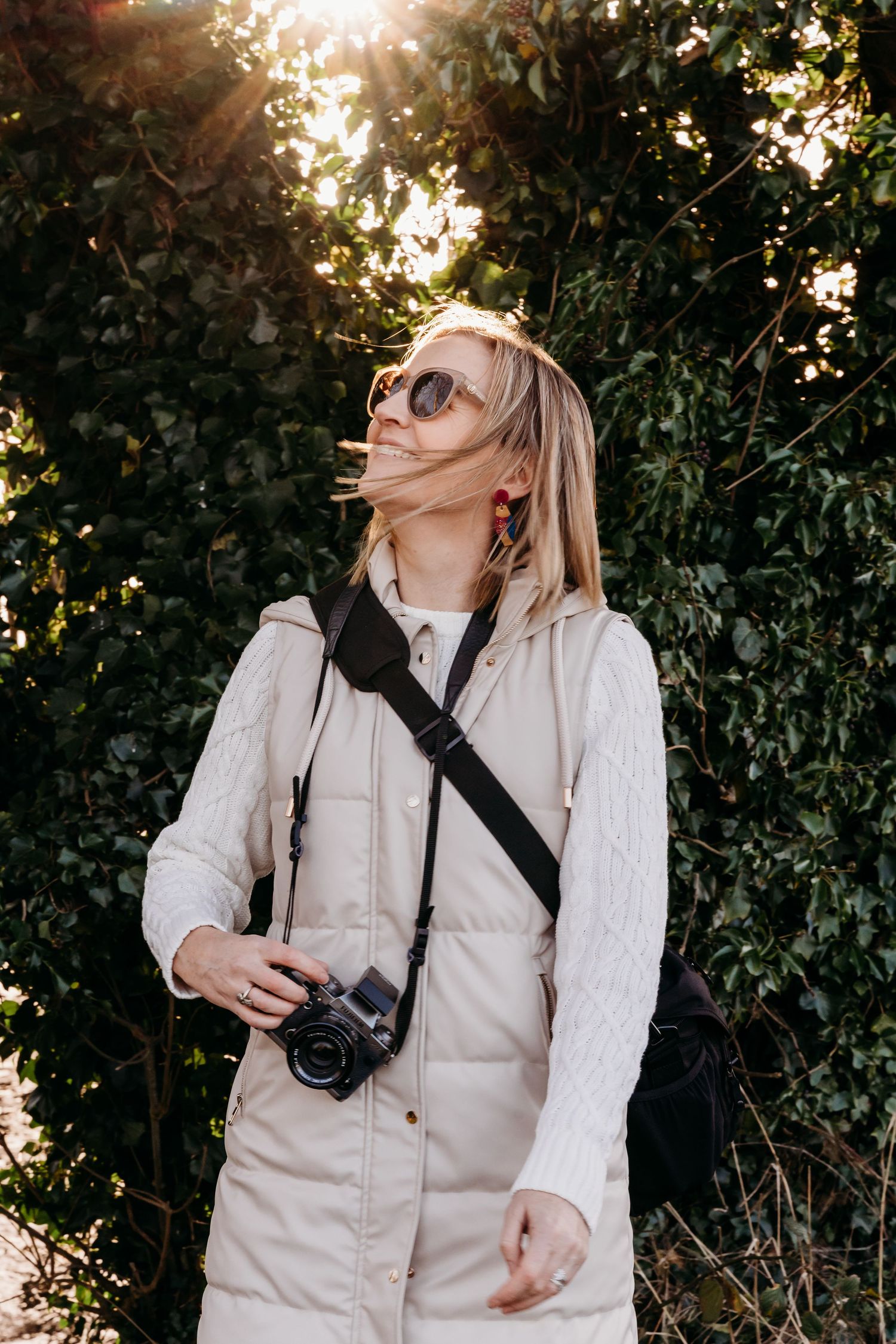 Photographer in light-colored vest and sunglasses smiles while carrying camera with leafy green backdrop at sunset.
