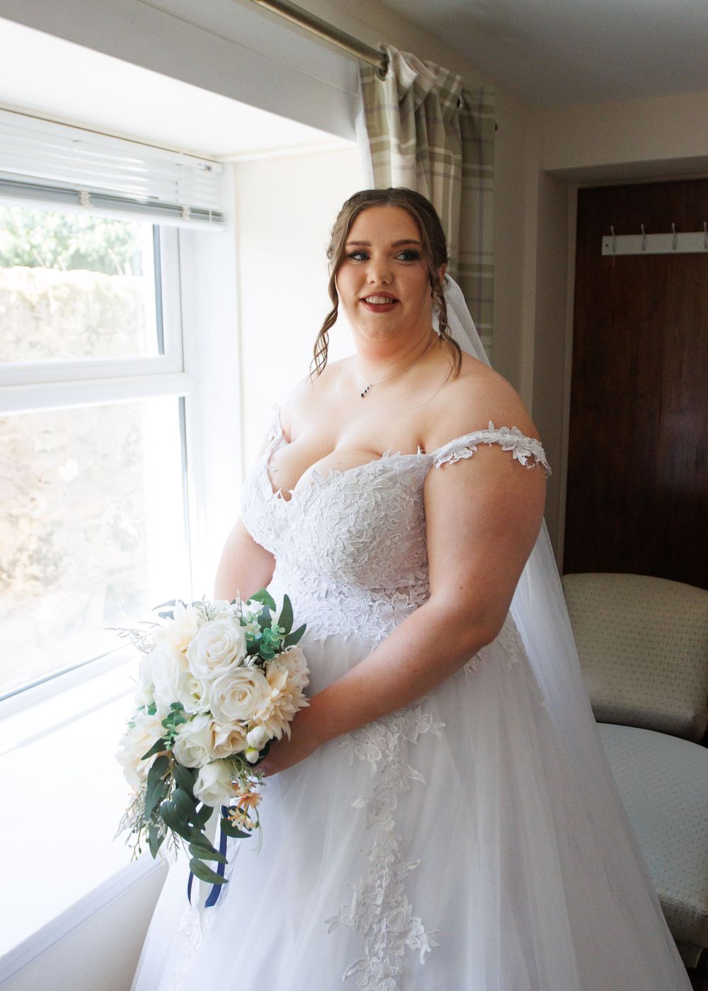 A bride in an elegant white lace gown holds a classic white and green bouquet by a sunlit window.
