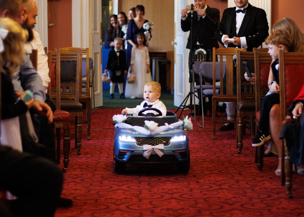 A young child rides a small blue toy car down a red-carpeted aisle during a formal event.