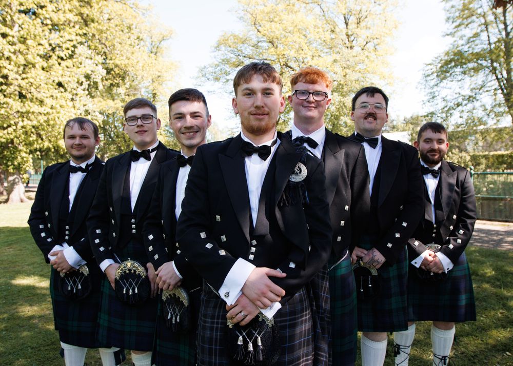 A group of groomsmen in traditional Scottish kilts and formal attire pose outdoors during a wedding celebration.