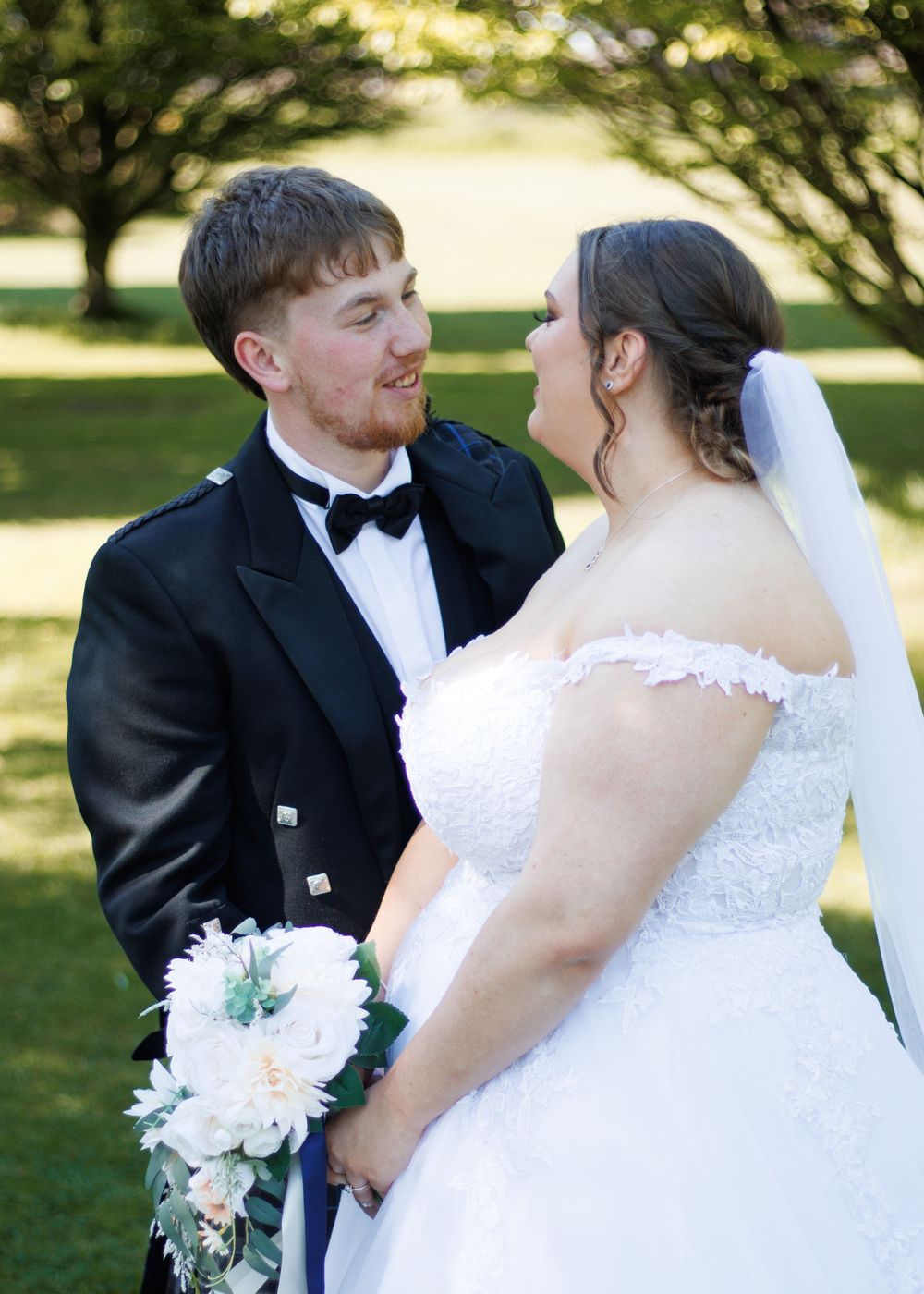 A couple shares a tender moment at their outdoor wedding, with the person wearing a black tux and kilt and white gown smiling at each other.