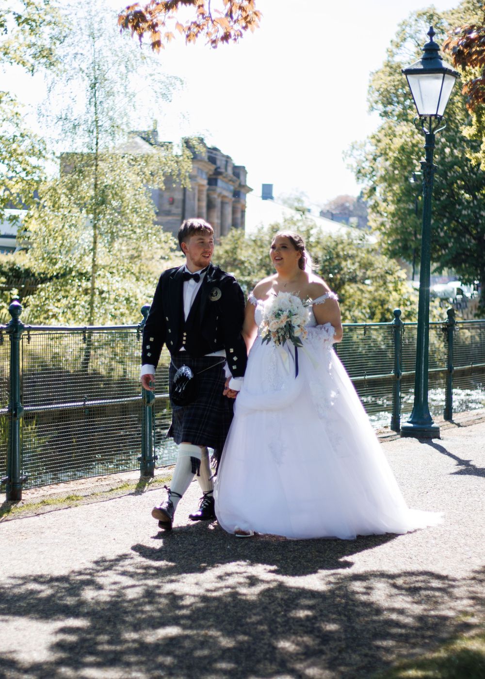 Traditional Scottish wedding couple posing outdoors on a path near lamppost with autumn trees in background.