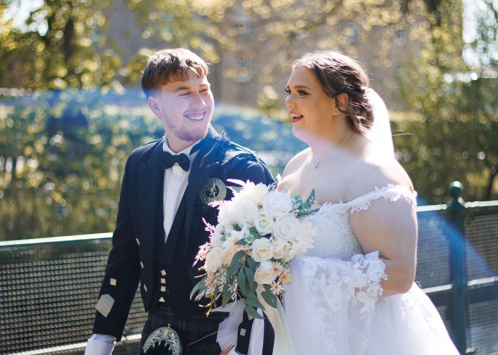 A bride holding white flowers and groom in a kilt pose together outdoors on a sunny day.