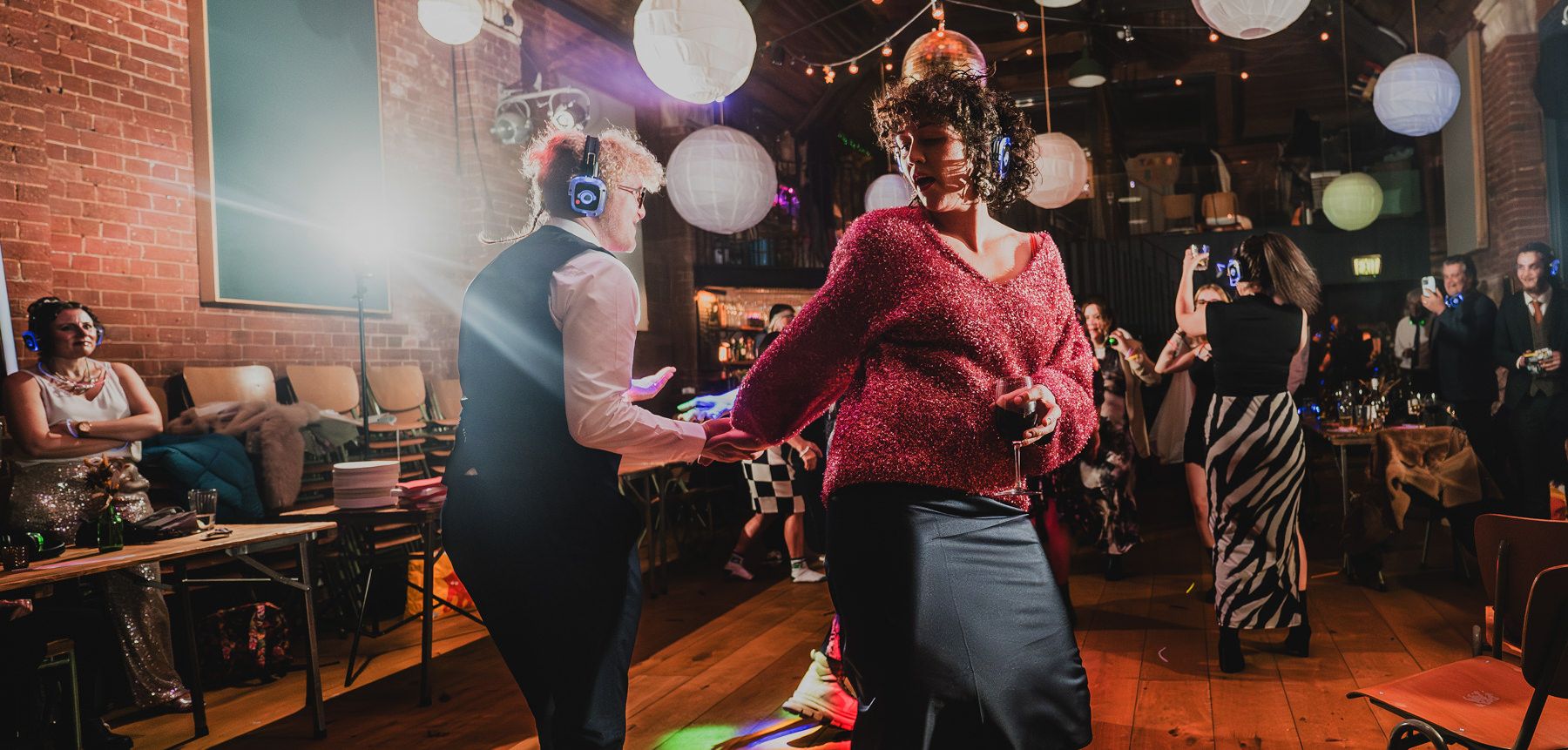 People dancing at a festive indoor celebration with colorful balloon decorations and ambient lighting.