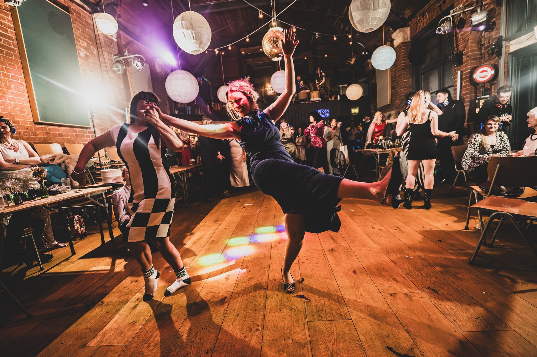 Wedding guests dance together on a wooden floor under glowing paper lanterns at an evening reception party.