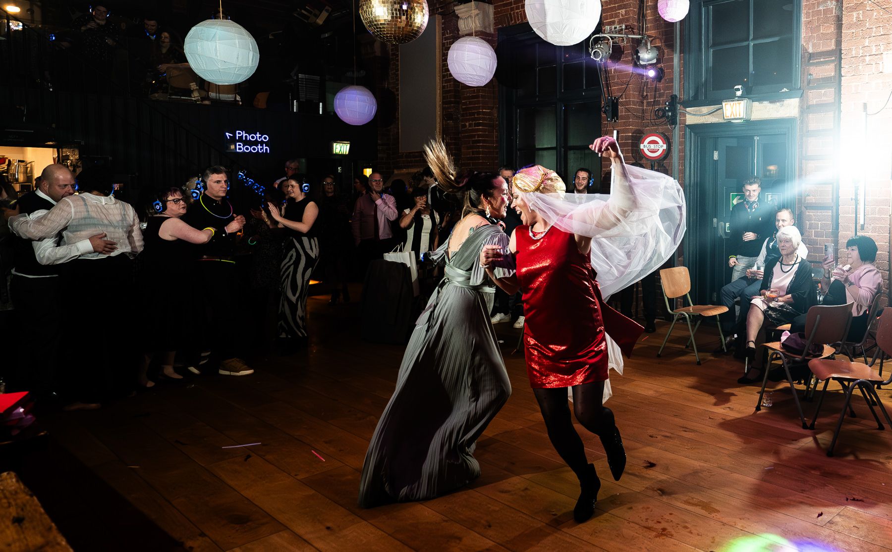Several dancers in motion under round paper lanterns on a hardwood floor with spectators in the background.