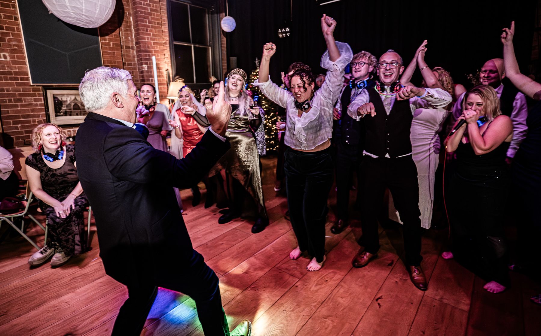 Energetic crowd dancing and celebrating with raised hands on a dance floor illuminated by pink and purple lighting.