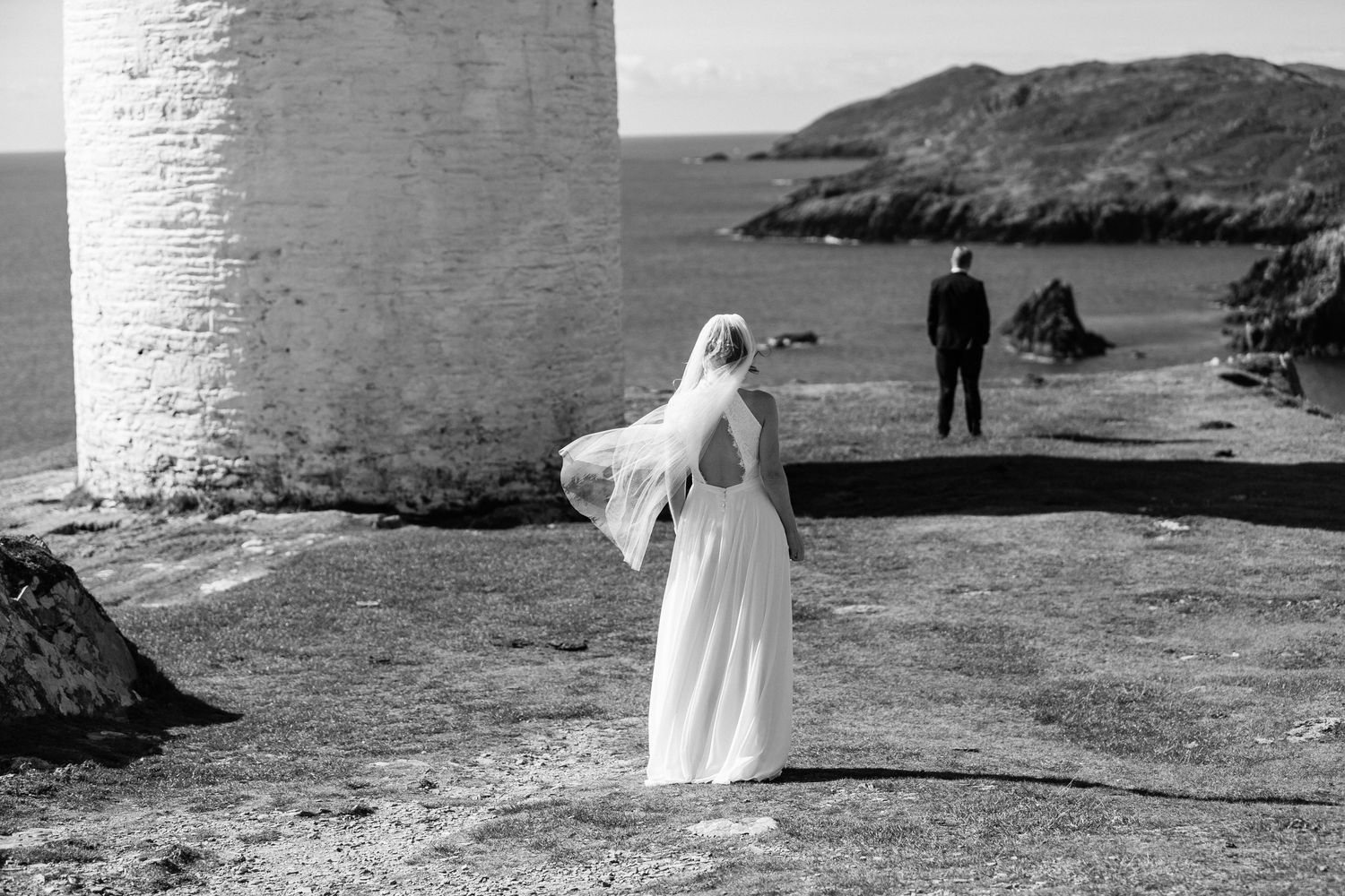 A couple walks along a scenic coastal cliff path with white lighthouse and rugged ocean views in monochrome.