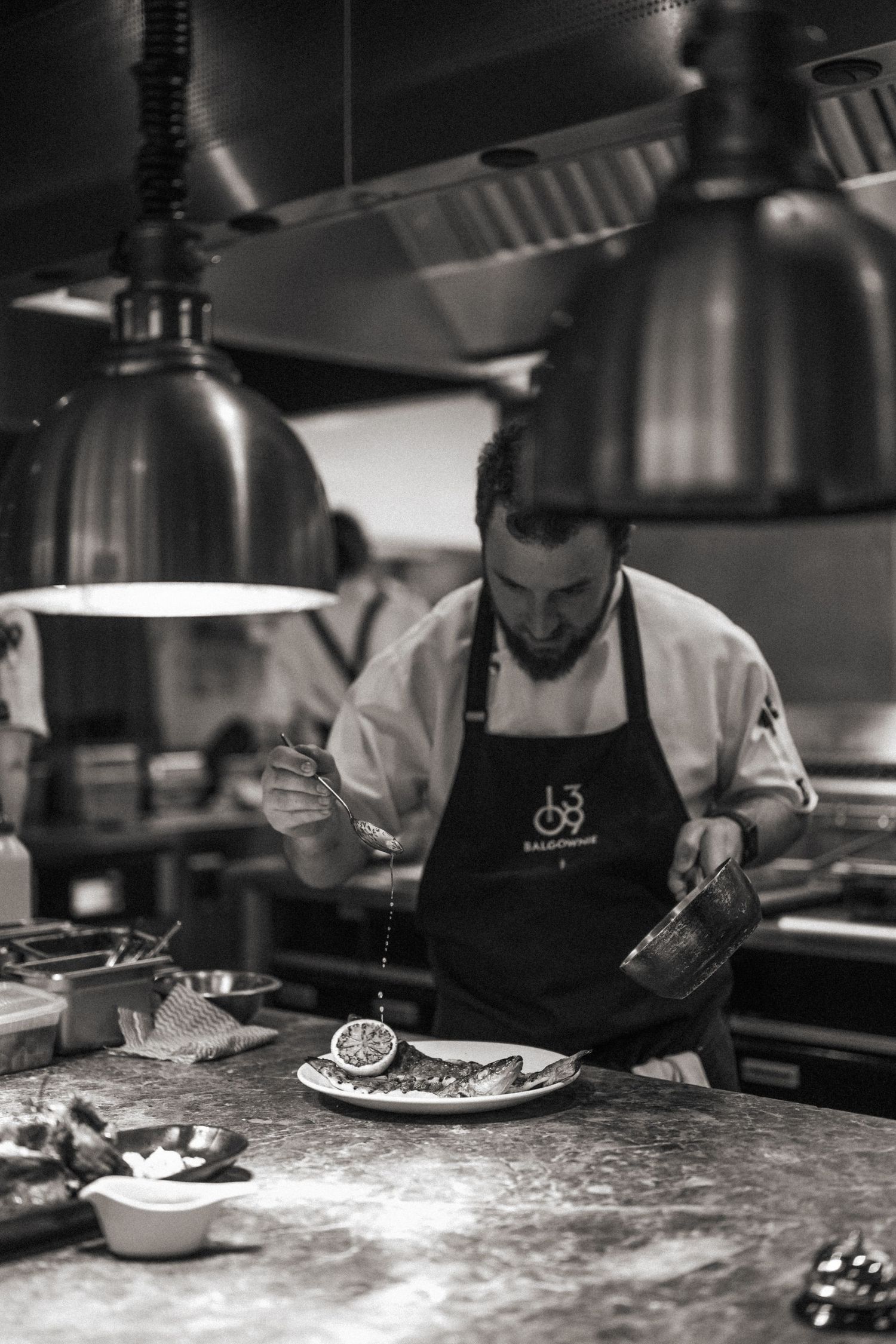 Professional chef in black apron carefully plating a fish dish with precise garnishing technique in a restaurant kitchen