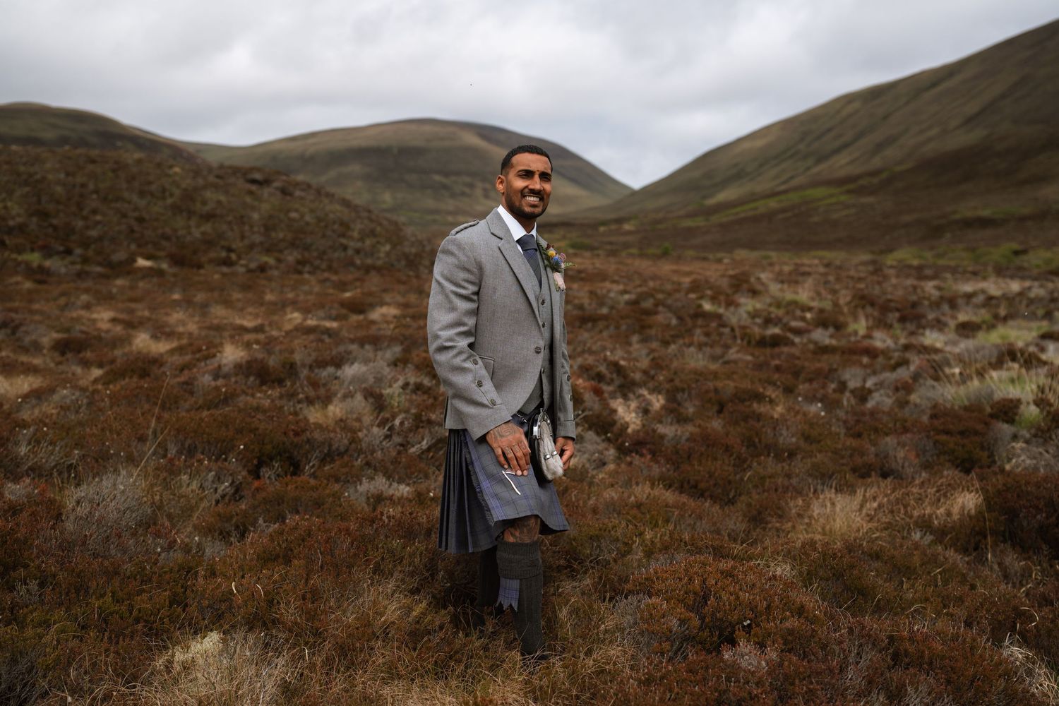 Someone wearing a grey suit stands in a rugged Scottish Highland landscape with heather and rolling hills on an overcast day.