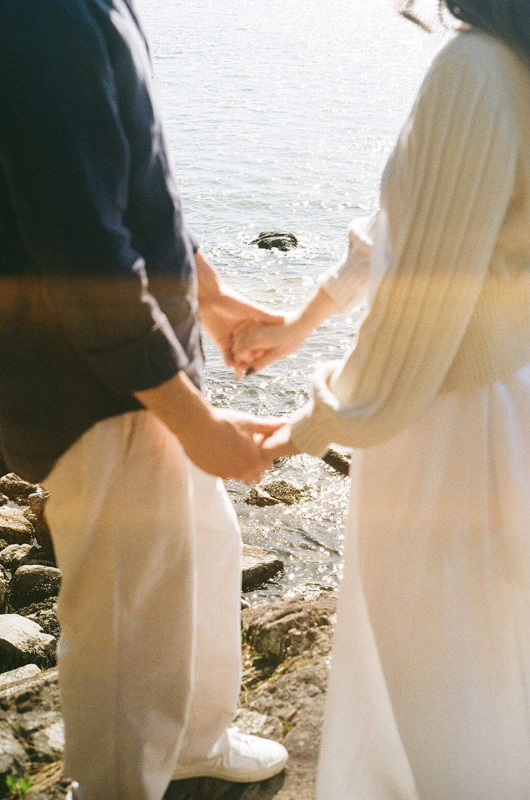 Close-up shot of a couple holding hands on a rocky beach with white fabric flowing in the wind.