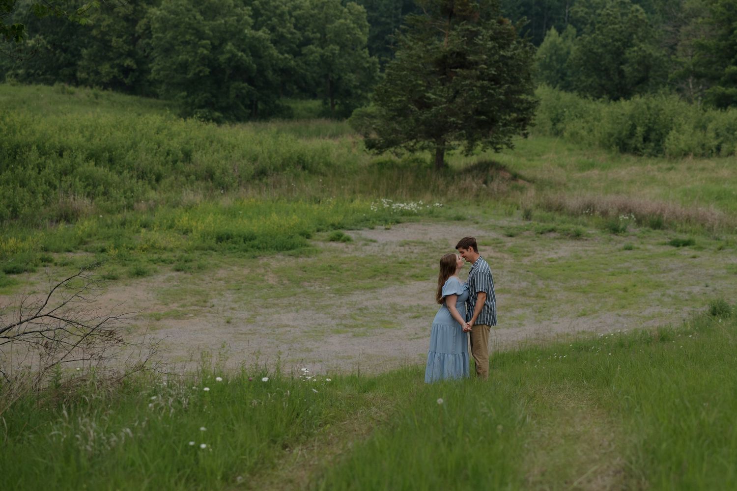 A couple in summer attire stands together on a dirt path surrounded by green meadows and trees.