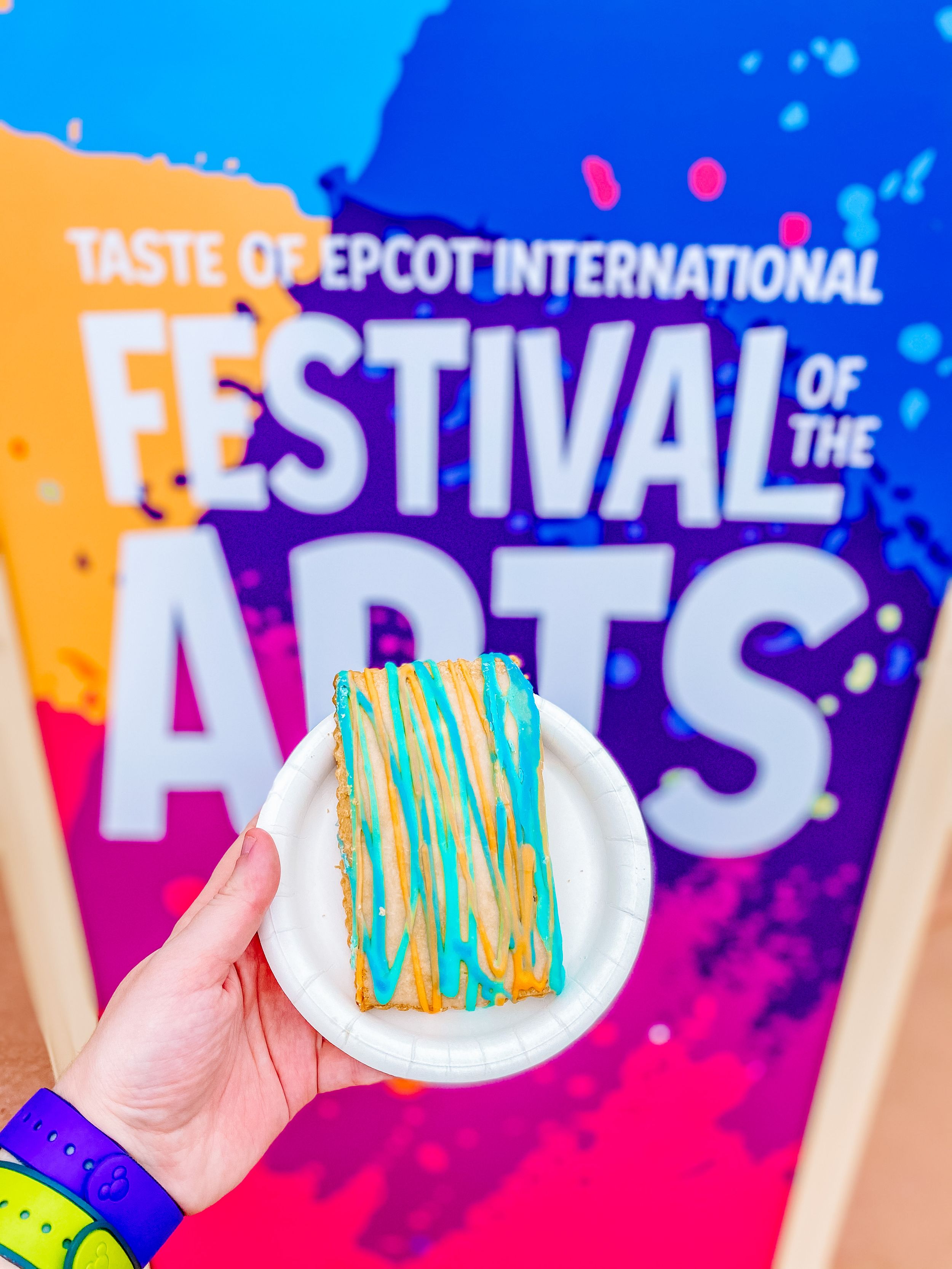 Turquoise and white decorated cookie on plate against colorful Festival of the Arts backdrop.
