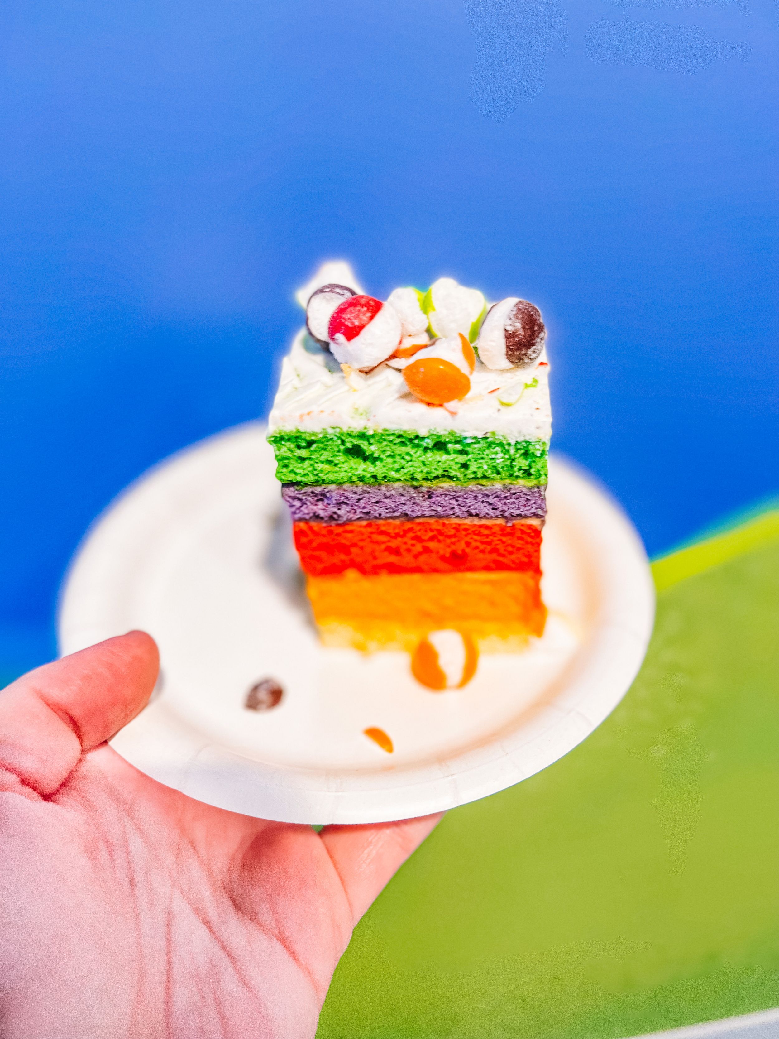 Layered rainbow cake slice with white frosting on white plate against blue background.