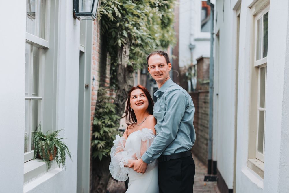 A couple stands close together in a charming narrow alleyway between white buildings in Utrecht with greenery visible.