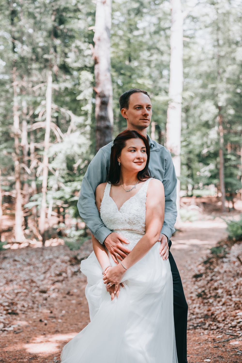 A couple embraces in a serene forest setting, the bride wearing a white lace gown while surrounded by tall evergreen trees.