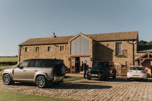 Modern cars parked outside a rustic stone barn conversion with large glass windows and cobblestone driveway.