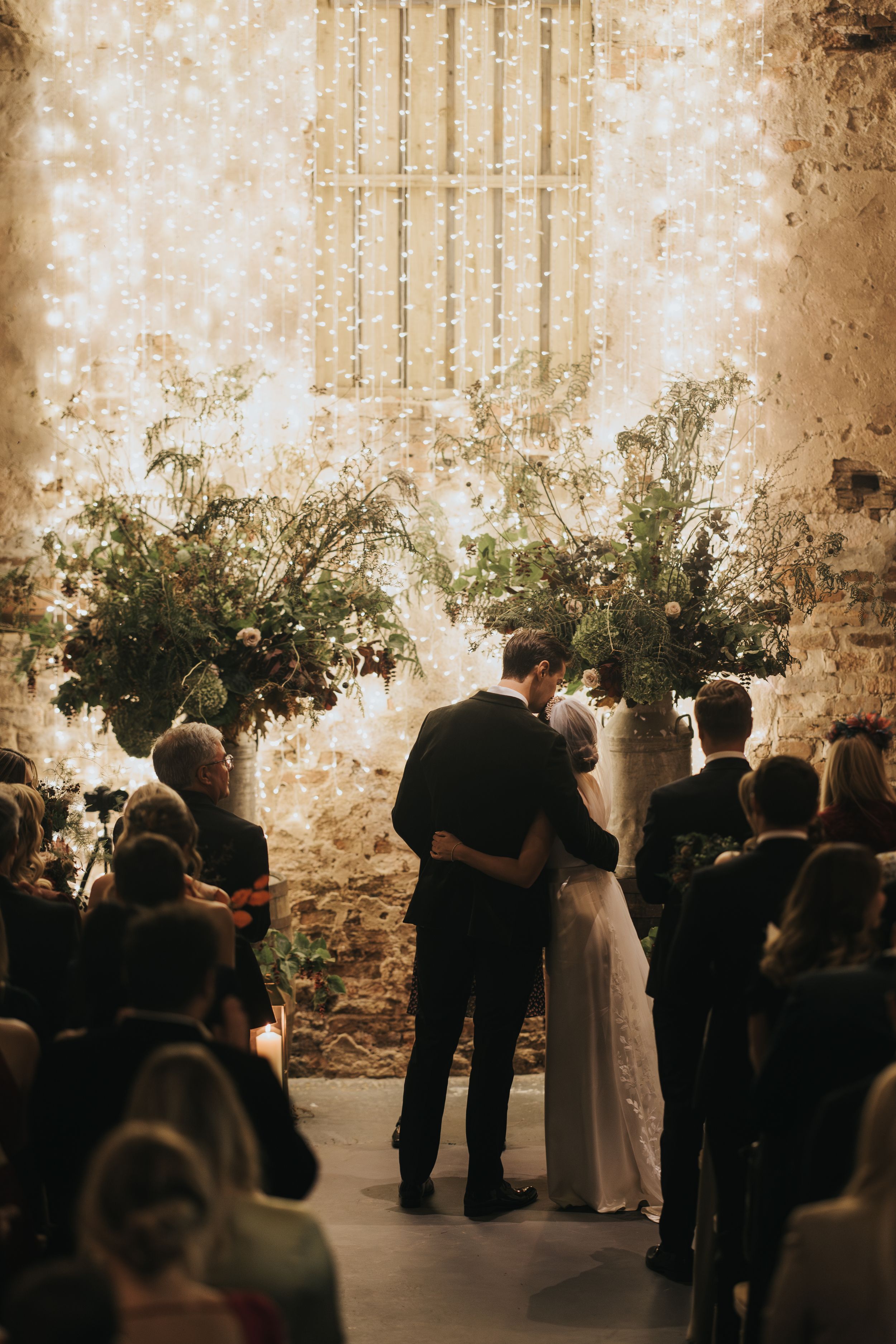 A wedding ceremony takes place in a rustic venue with stone walls and lush greenery arrangements in a warm sepia tone.