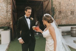 A couple in formal wedding attire share a moment in a brick-walled venue with natural lighting.