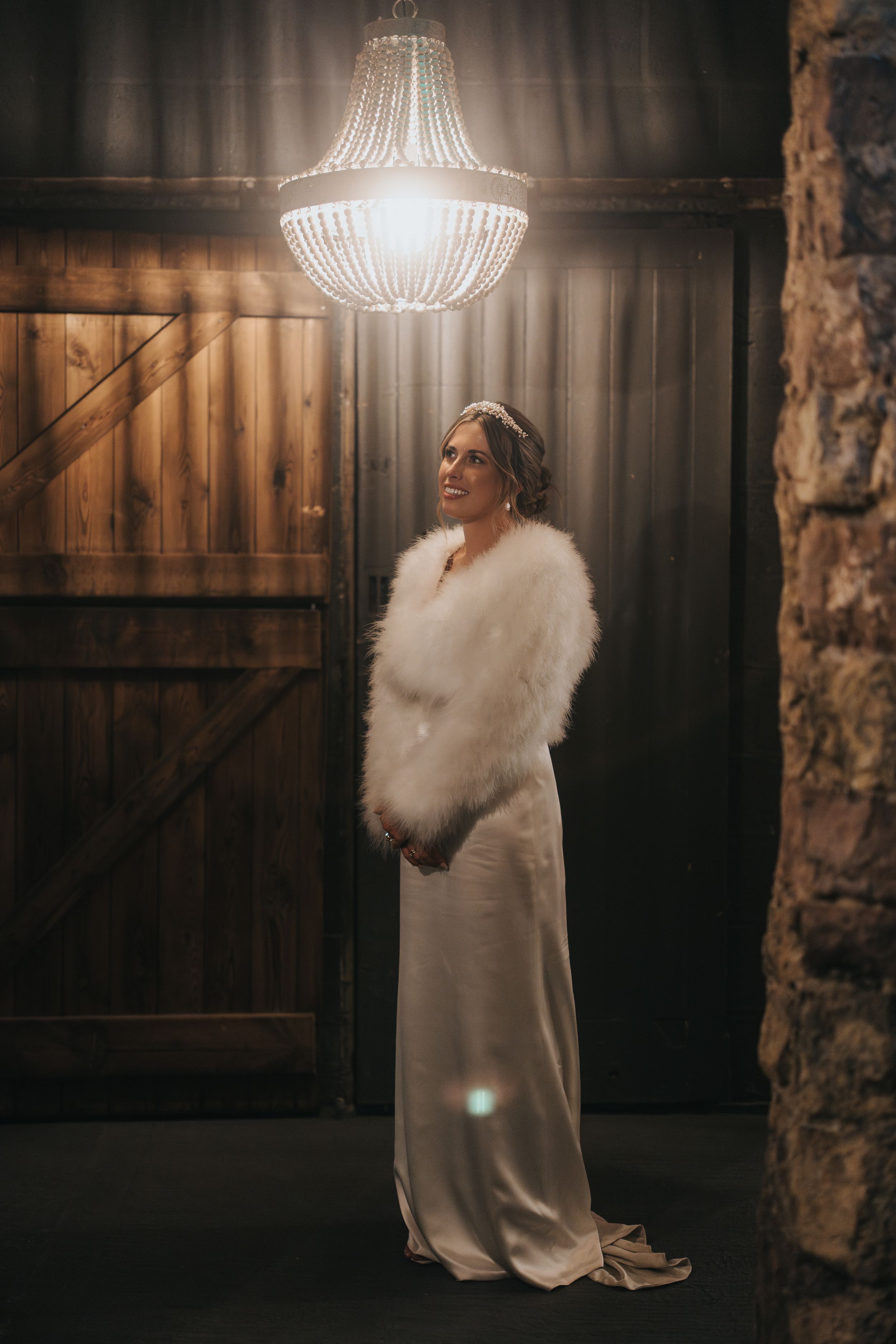Elegant bride in white gown and fur stole poses beneath crystal chandelier in rustic barn setting.