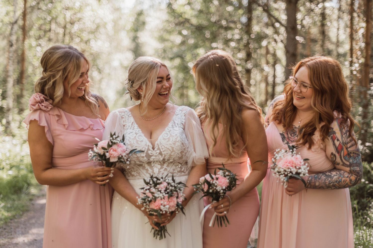 Bridal party in blush pink and white dresses pose with floral bouquets in a sunlit forest setting.