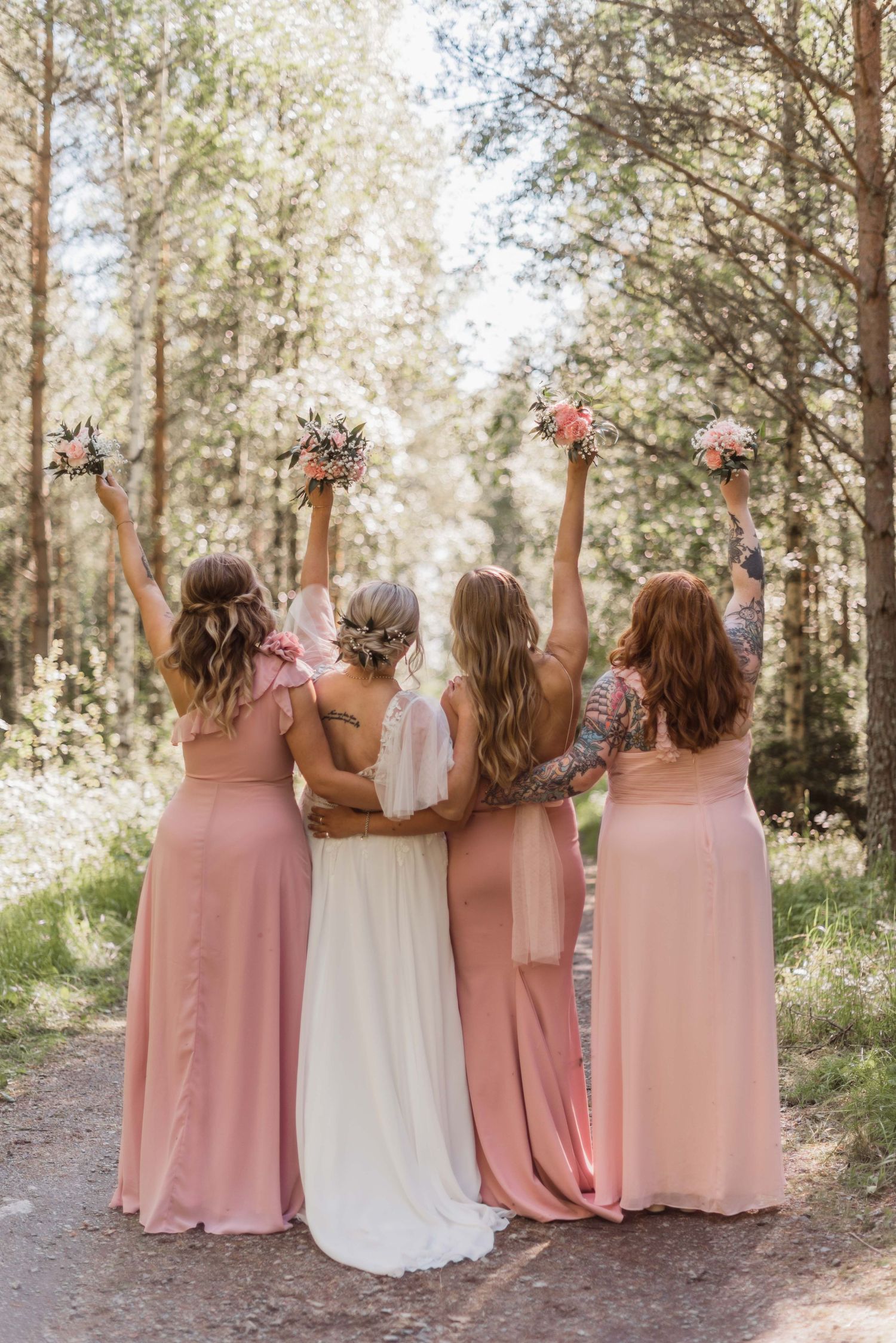 A wedding party in pink bridesmaid dresses and white wedding gown raise their bouquets together in a sunlit forest.
